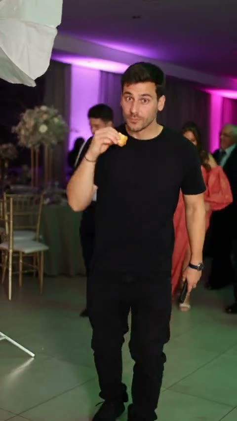 Frame from the Bby Wow meme clip: a man in a black t-shirt holding a snack to his mouth and staring at the camera in a purple-lit banquet hall
