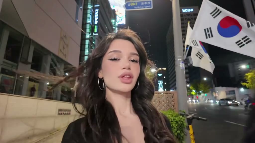 Frame from the Gangnam Style TikTok trend: a woman filming a selfie video on a Seoul street at night under Korean flags