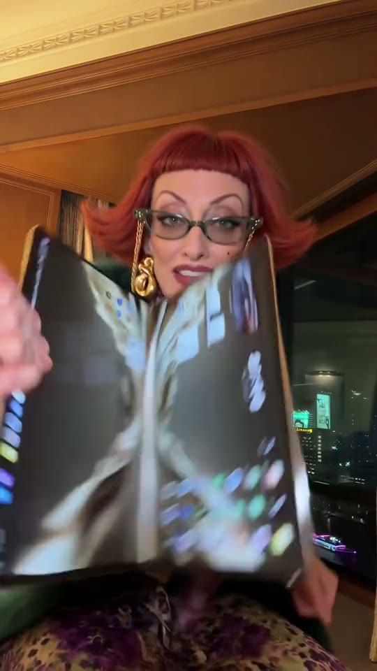 iPhone Kupi 热梗视频的一帧：红发、戴猫眼眼镜的女士在酒店房间举着正在展开的手机