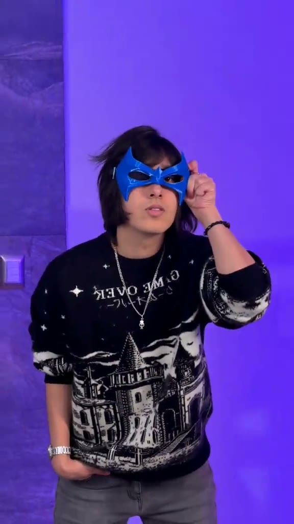 Frame from the Nightwing TikTok trend: a man in a black graphic sweater holding a blue Nightwing mask to his face