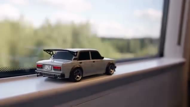 Frame from the toy car to real car clip: a white Lada scale model with a rear wing on a windowsill, trees outside the window