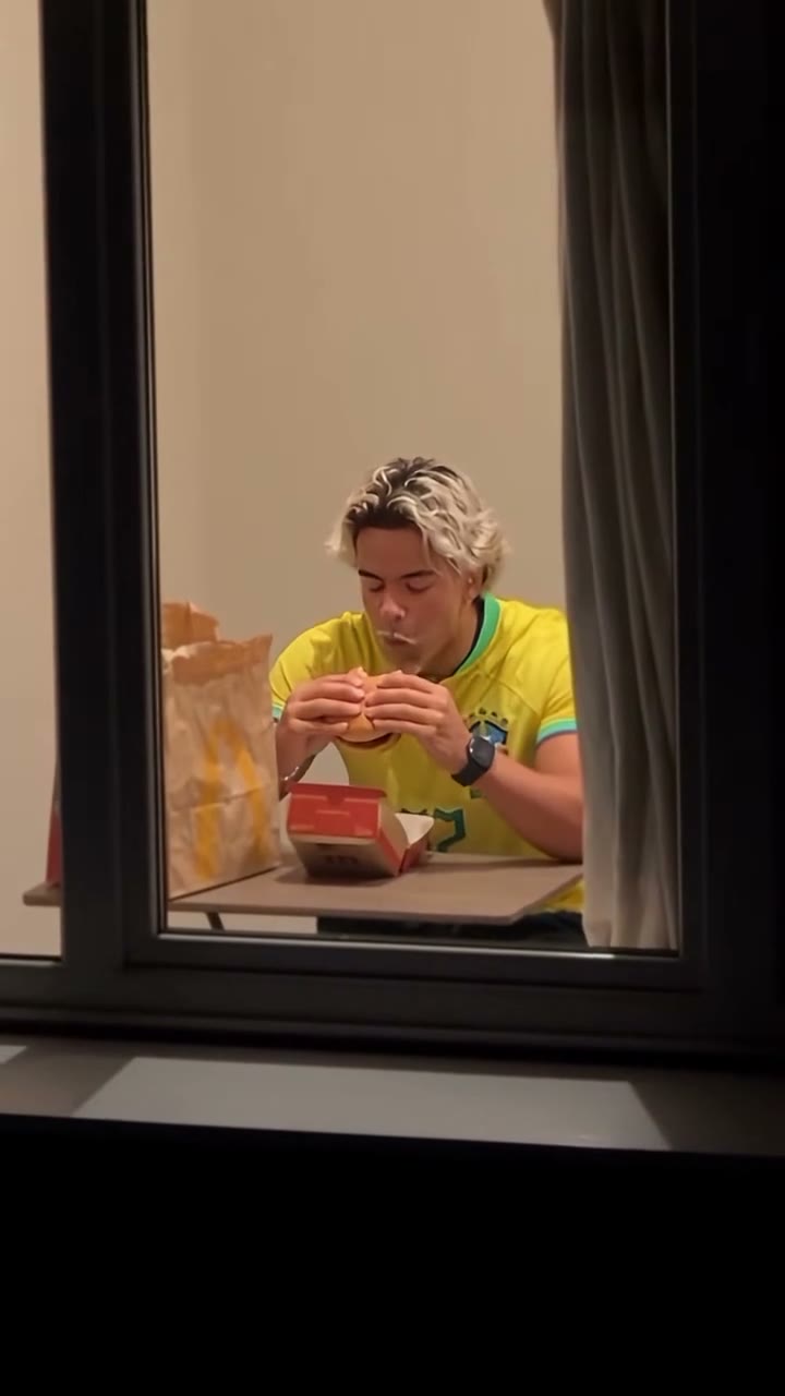 Frame from the window peeper TikTok trend: a man in a yellow football shirt eating a burger behind a lit apartment window at night